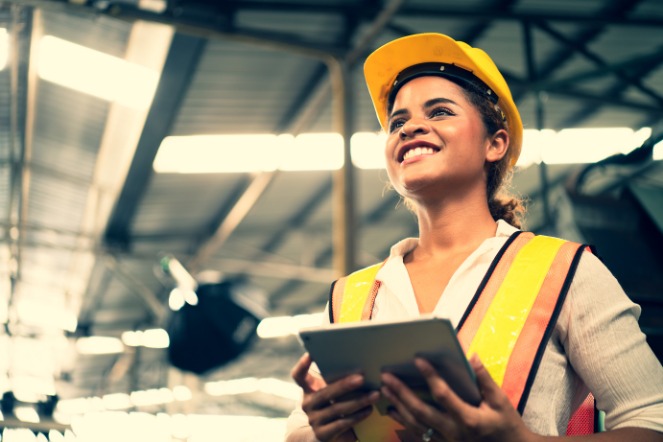 A mechanical design engineer holding a clipboard standing in a warehouse looking very proud of the amazing work she has completed